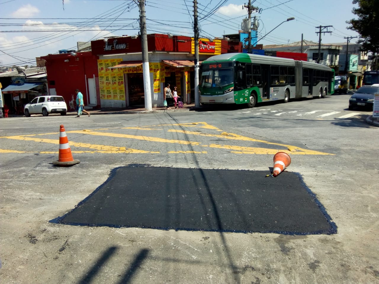 Retângulo preto no centro da rua Douradoquara, é a marca do asfalto refeito. Houve a compactação e alinhamento com a parte que não foi afetada. Ao fundo, um ônibus articulado está parado no ponto.